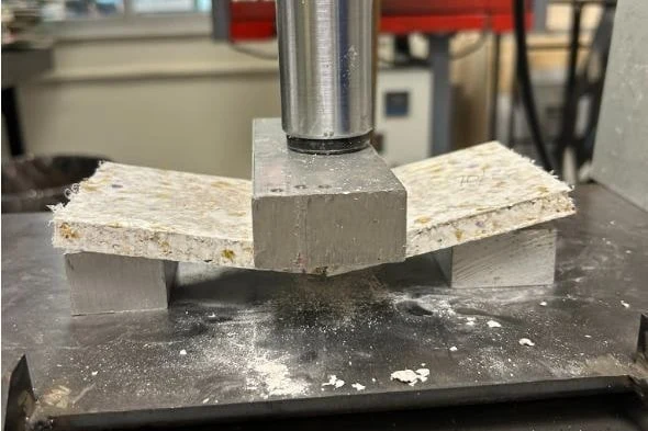 A composite tile snapped into a shallow V under the head of a hydraulic press, crushed shell visible along the fracture.
