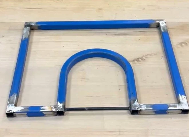 A welded steel pressing frame on a workbench — a rectangle with an arc bridging its base, the paint sandblasted back to bright metal at every weld.