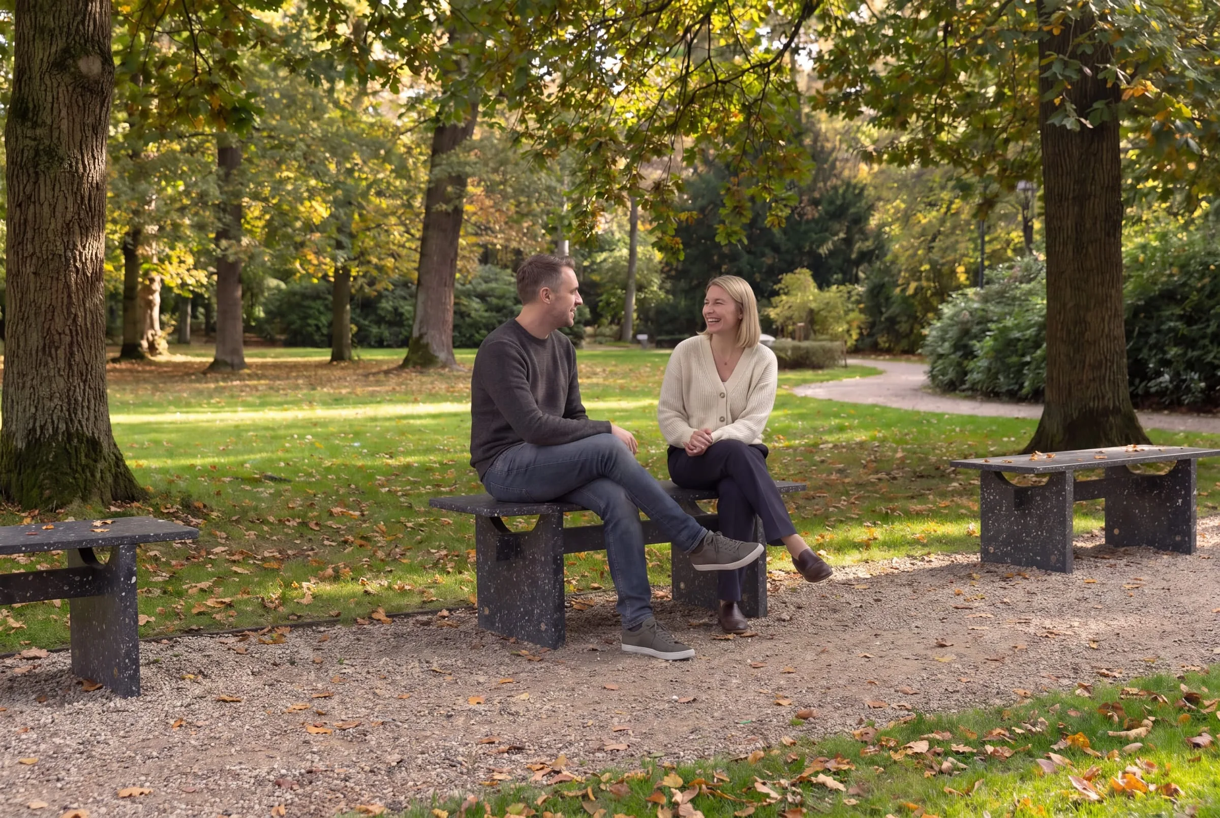 Two people sitting and talking on a Shio bench in a park, with more benches along the path behind them.
