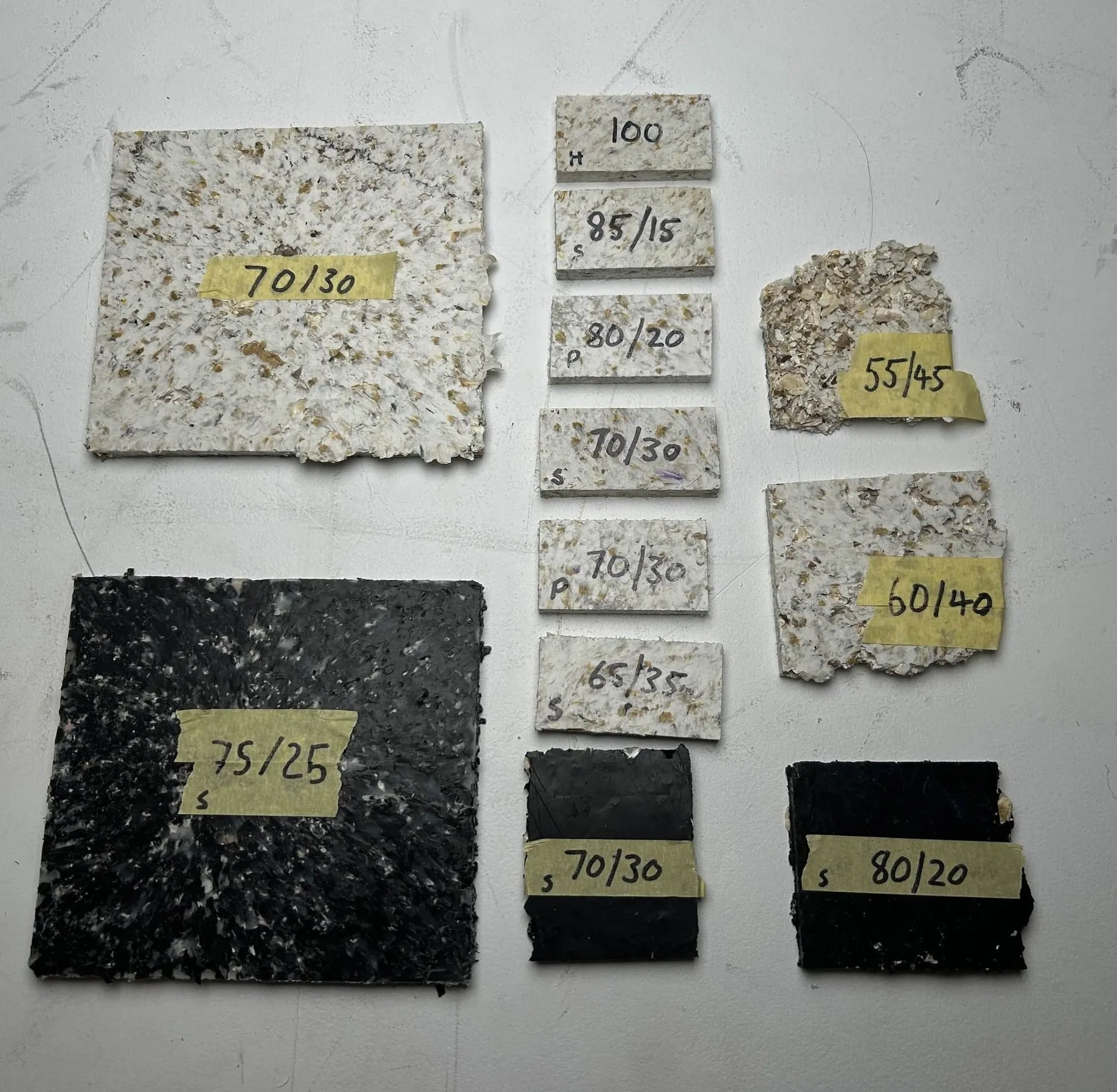 Twelve composite test tiles laid out on a concrete floor, each labelled in marker with its HDPE-to-shell ratio — 100, 85/15, 80/20, 75/25, 70/30, 65/35, 60/40, 55/45.
